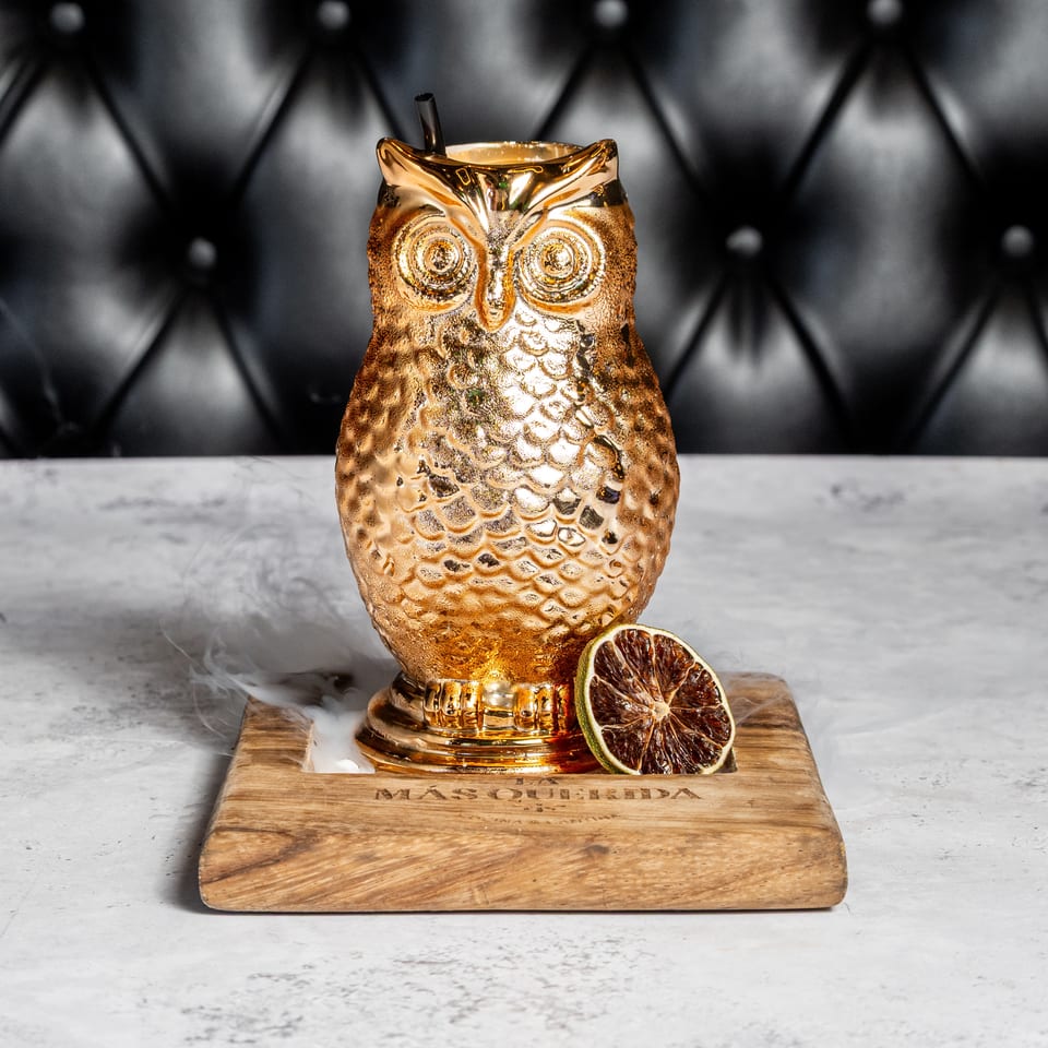 Golden owl-shaped cocktail vessel with a straw, dried citrus garnish, and swirling smoke on a wooden board.