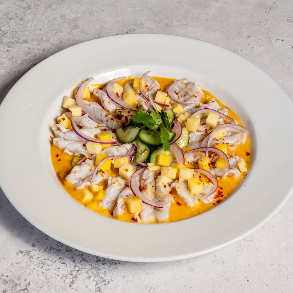 Ceviche-style seafood dish with mango, cucumber, red onion, cilantro, and chili flakes in orange sauce on a white plate.