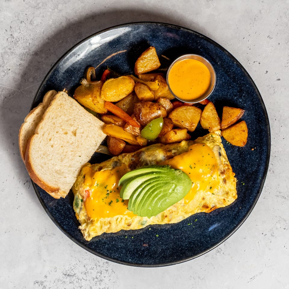 Cheesy avocado-topped omelet served with toasted bread, roasted potatoes and vegetables, and a side of orange sauce.