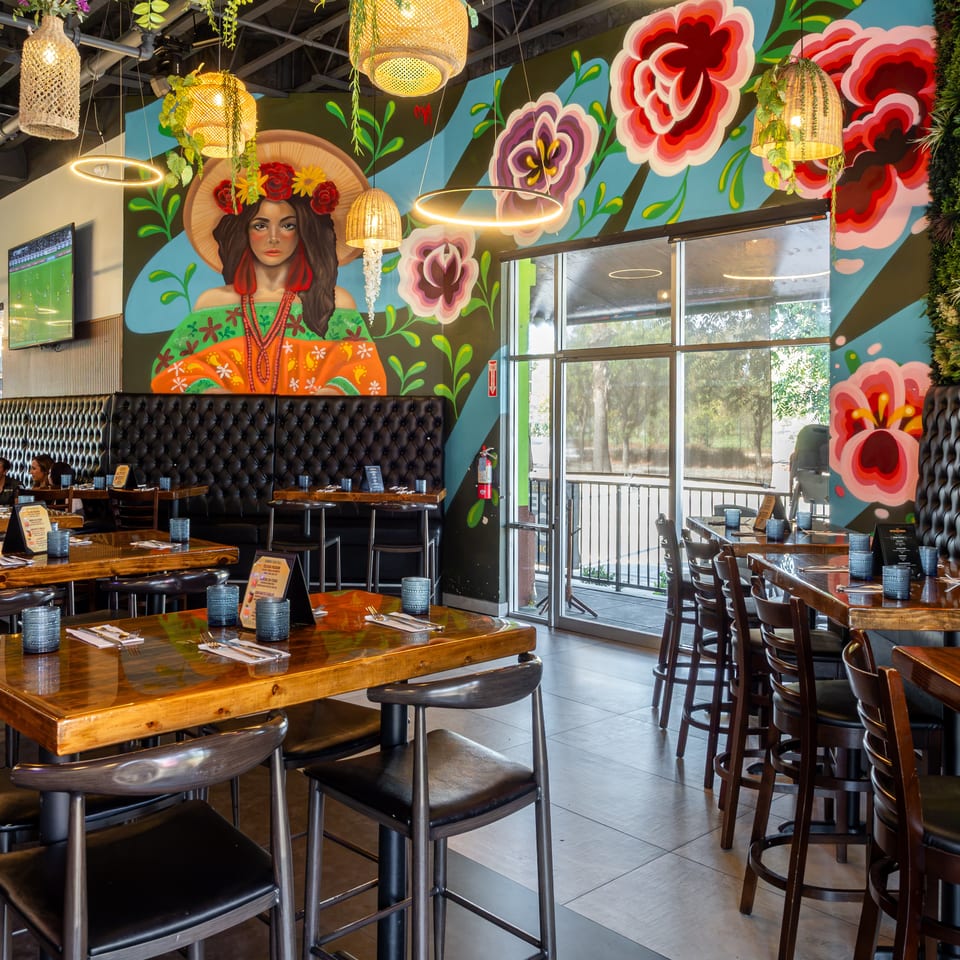 Vibrant restaurant dining room with floral mural, portrait of a woman, hanging wicker lights, wooden tables, and tufted booths.