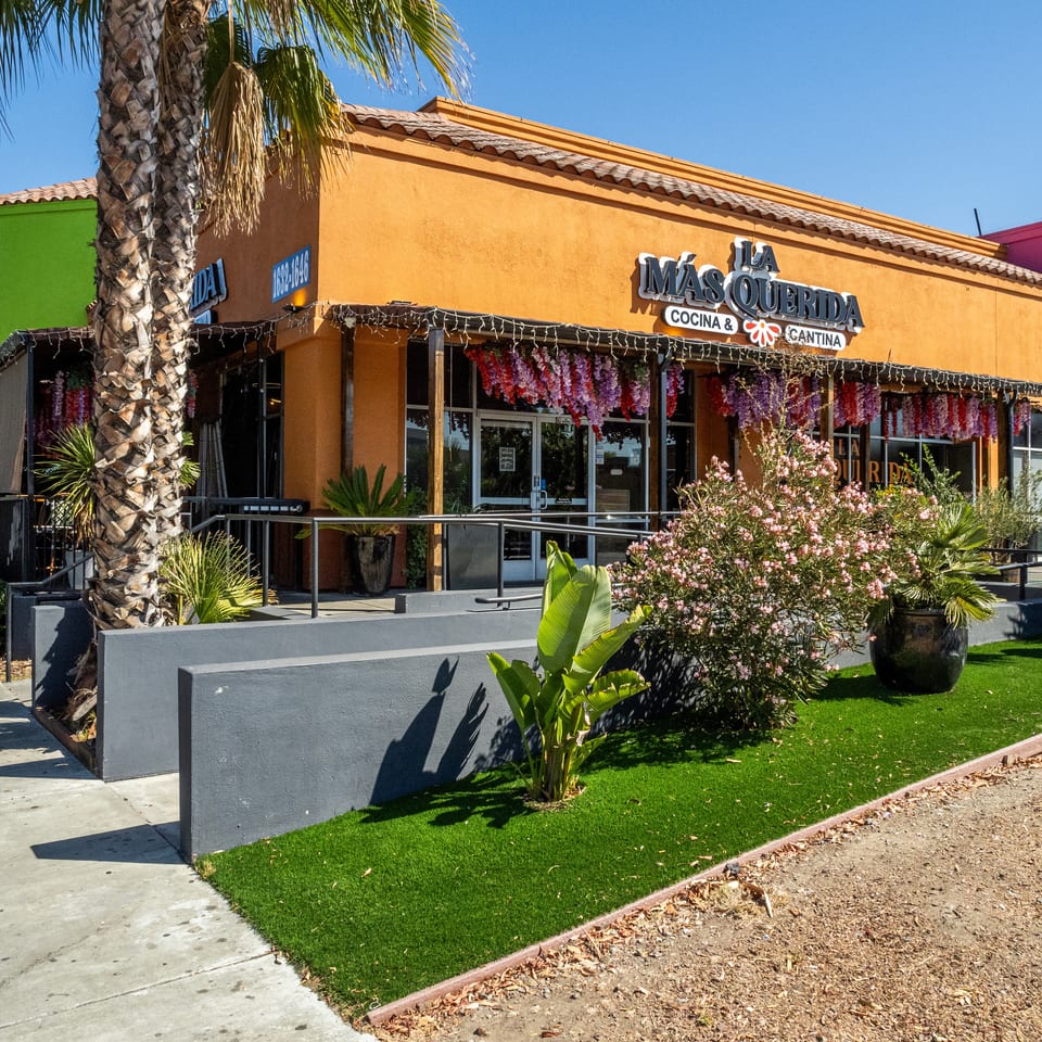 Exterior of La Más Querida Cocina & Cantina, a colorful Mexican restaurant surrounded by palm trees and flowering plants.
