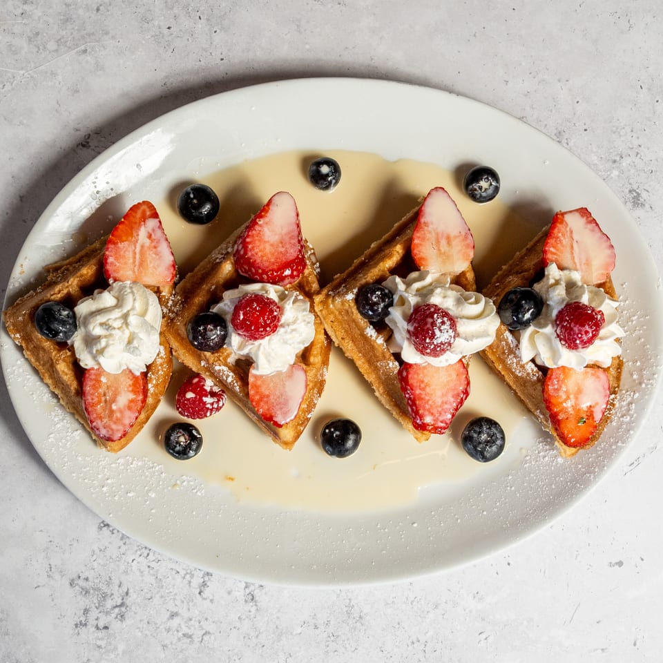 Four golden waffles topped with whipped cream, sliced strawberries, raspberries, blueberries, powdered sugar, and syrup on a white plate.