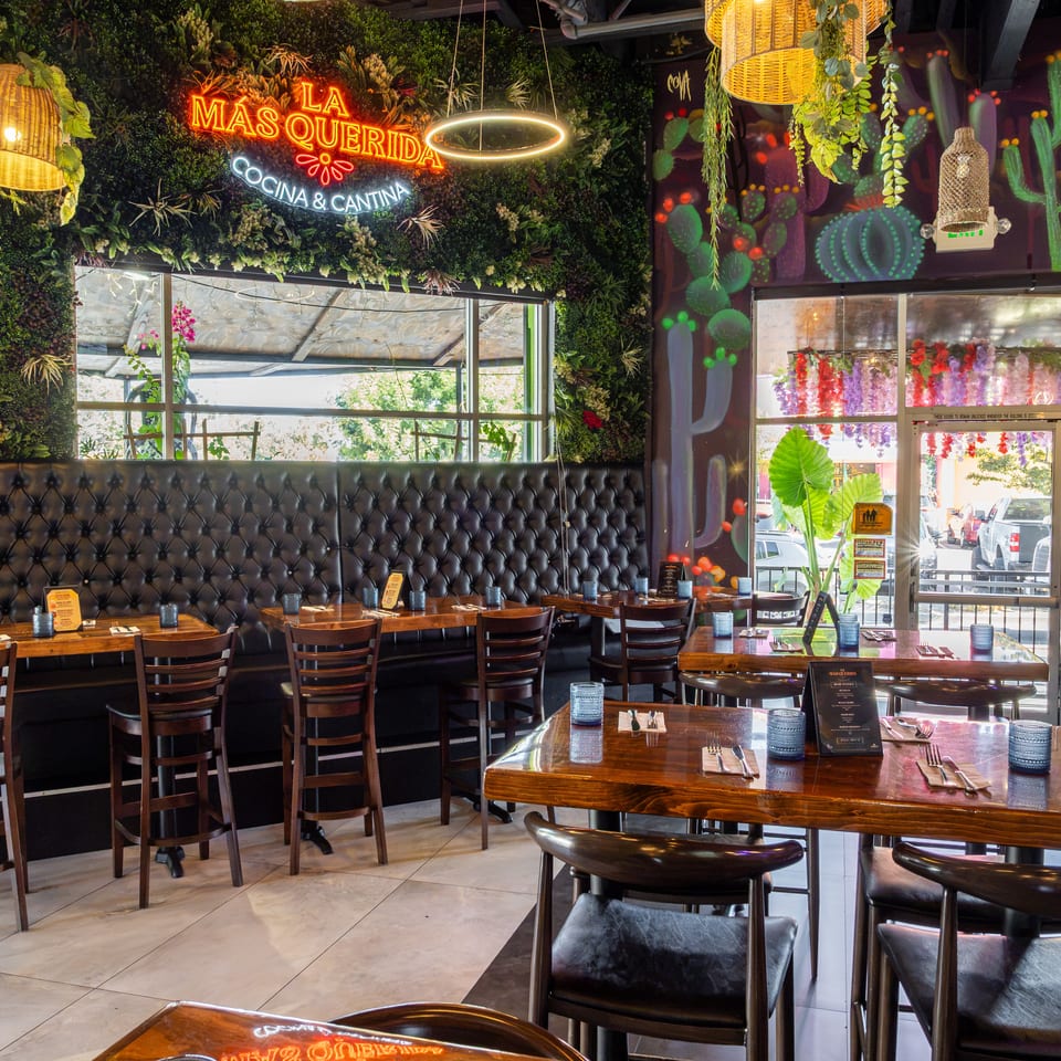 La Mas Querida Cocina & Cantina dining room with wooden tables, tufted seating, lush greenery, cactus murals, and vibrant hanging lights.