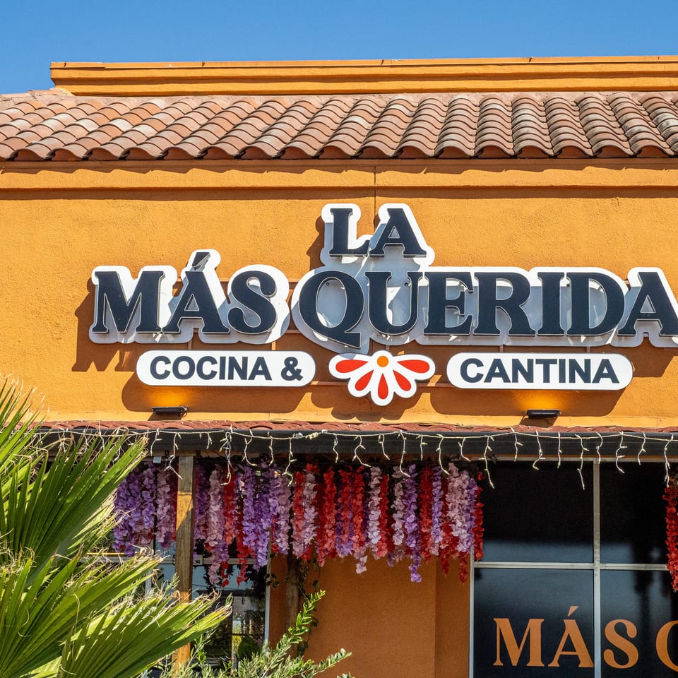 Exterior sign for Másquerida Cocina & Cantina on an orange stucco building with tiled roof, flowers, and palm leaves.
