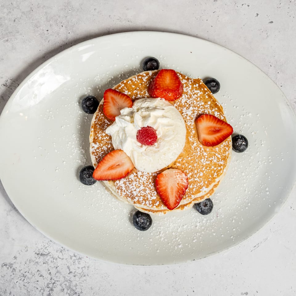 Stack of pancakes topped with whipped cream, strawberries, blueberries, raspberries, and powdered sugar on a white plate.