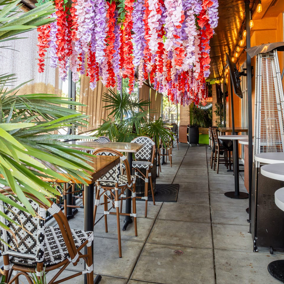 Colorful hanging flowers and tropical plants decorate an outdoor restaurant patio with patterned chairs, wooden tables, and patio heaters.