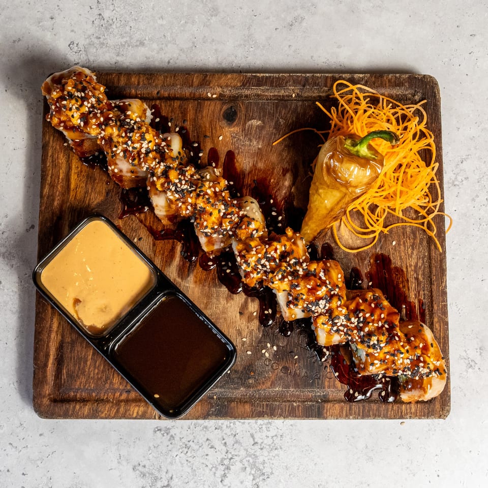 Sushi roll drizzled with spicy sauce and sesame seeds, served on a wooden board with dipping sauces, pickled ginger, and shredded carrot.