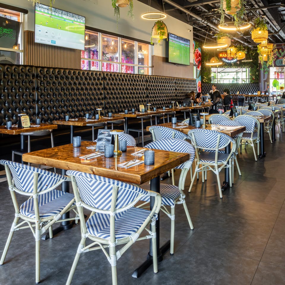 Colorful restaurant dining room with patterned chairs, wooden tables, tufted booths, hanging plants, pendant lights, and sports playing on televisions.