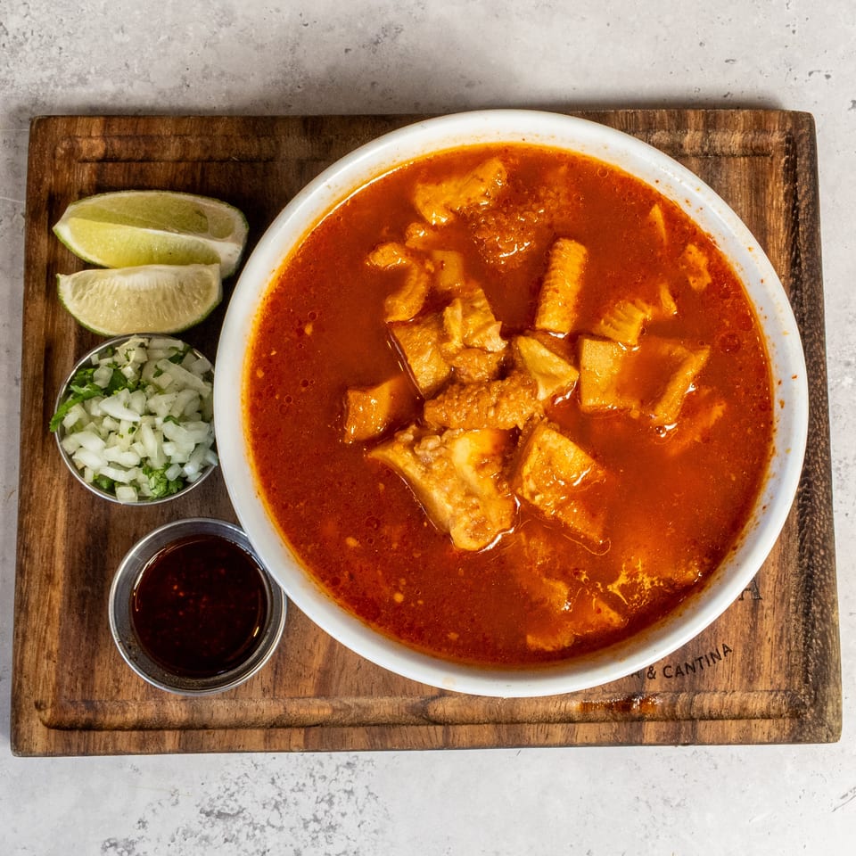 Bowl of rich red stew with chunks of meat, served with lime wedges, chopped onions, cilantro, and dark sauce on a wooden board.