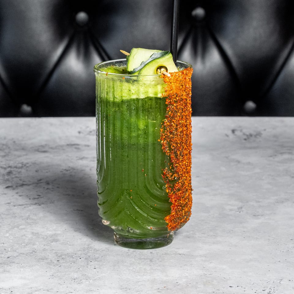 Green cocktail garnished with cucumber, herbs, and a chili-salt rim, served in a textured glass on a stone countertop.