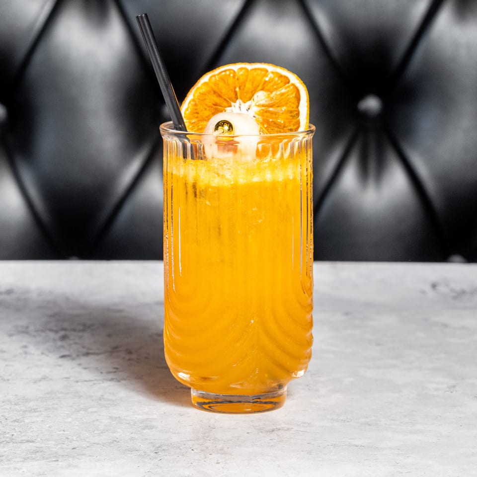 Bright orange cocktail in a textured glass, garnished with an orange slice and edible flower, sits on a marble bar.