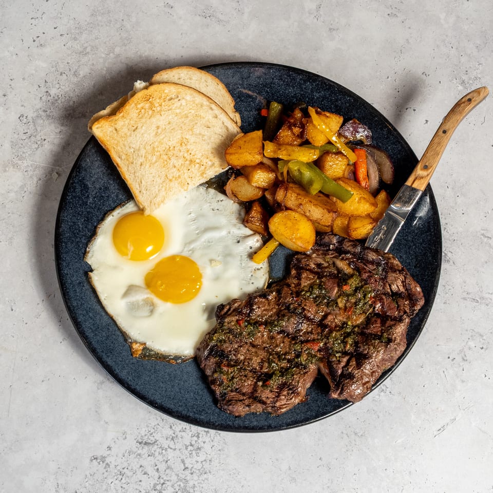 Grilled steak topped with chimichurri, two fried eggs, roasted potatoes and peppers, and toast on a black plate.