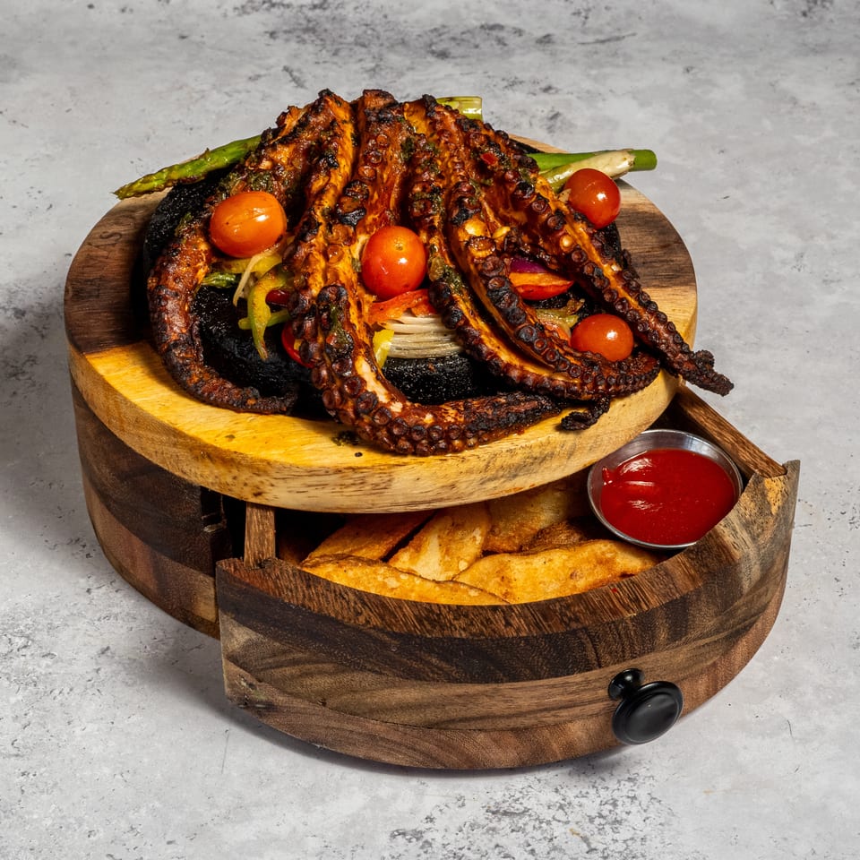Grilled octopus tentacles with vegetables and cherry tomatoes served on a wooden platter with fries and red dipping sauce.