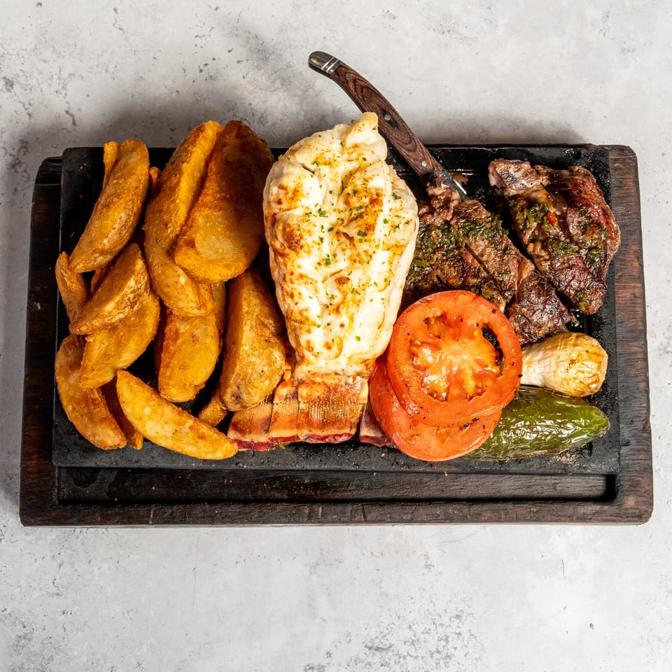 Grilled steak with herb sauce, cheesy baked potato, potato wedges, roasted tomato, pepper, and onion on a sizzling platter.