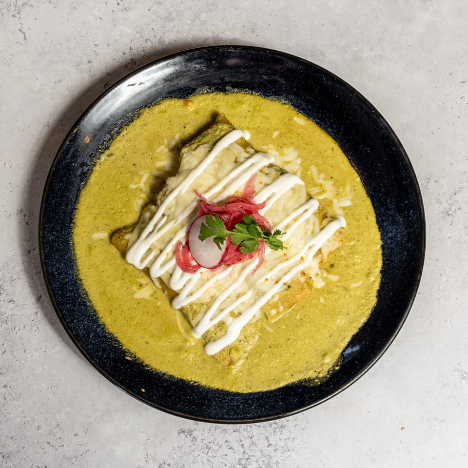 Black plate of enchiladas covered in green sauce, drizzled with crema and garnished with pickled onions, radish, and cilantro.