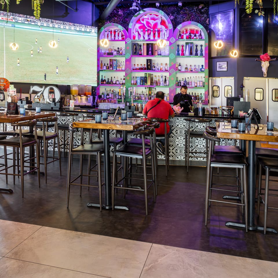 Vibrant restaurant bar with colorful liquor shelves, soccer match on a large screen, hanging plants, and high-top tables.