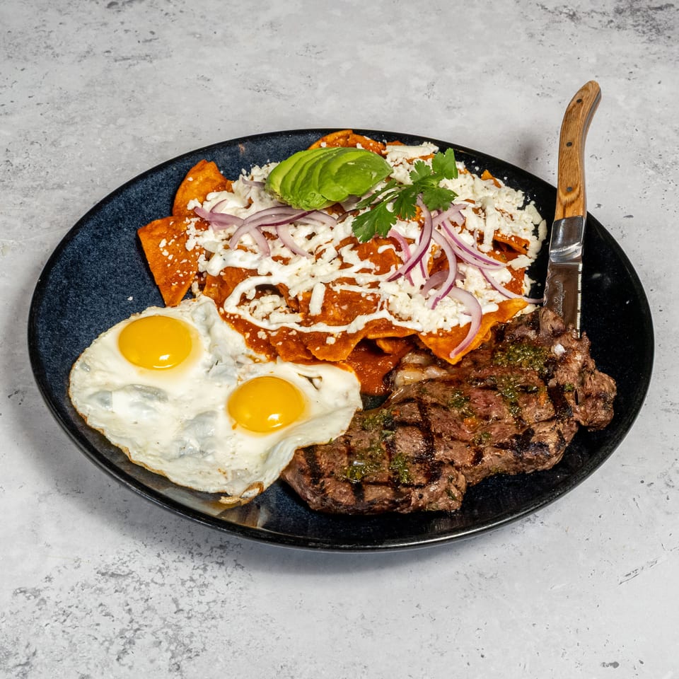 Mexican-style plate with red-sauced enchiladas, fried eggs, grilled steak, avocado, cheese, red onion, and cilantro.