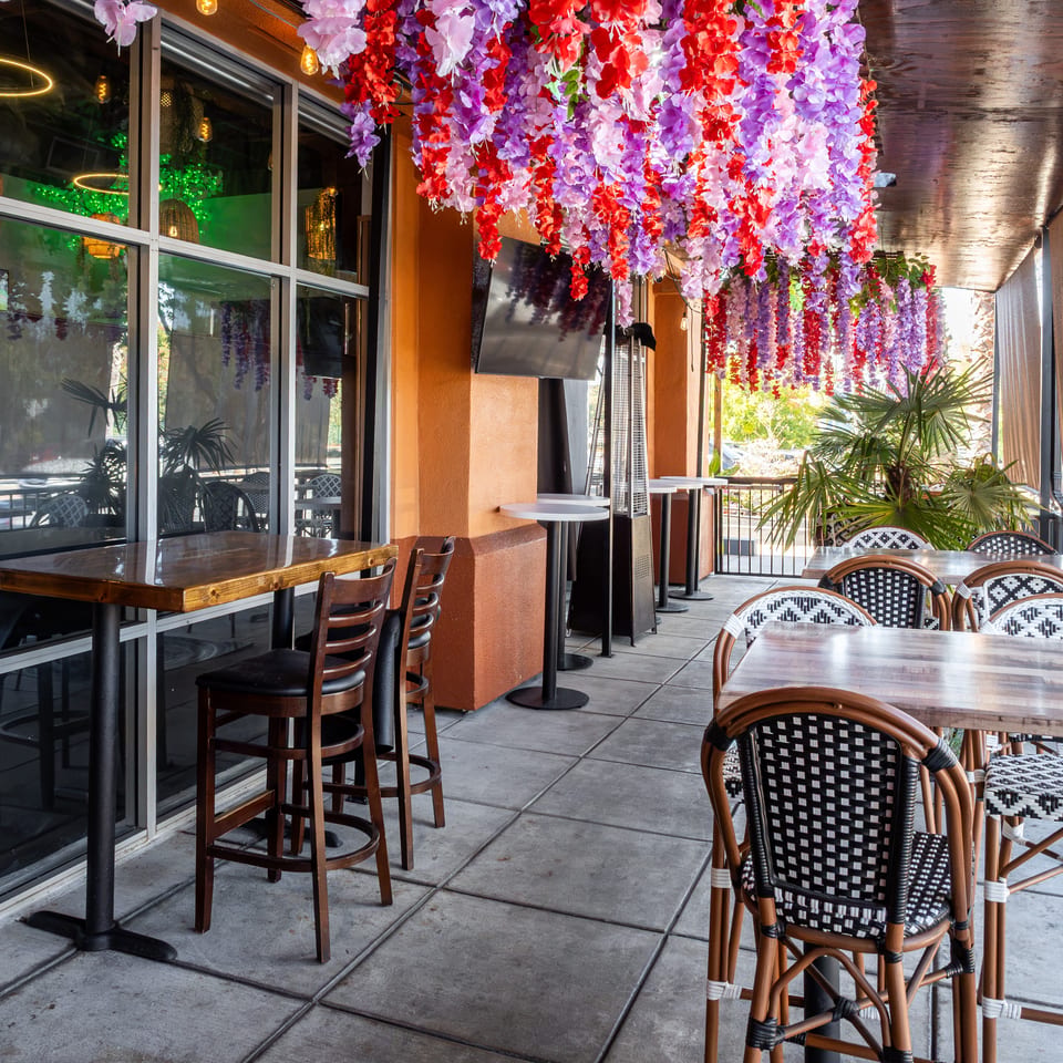 Restaurant patio with woven chairs, wooden tables, tropical plants, and colorful purple and red flowers hanging overhead.