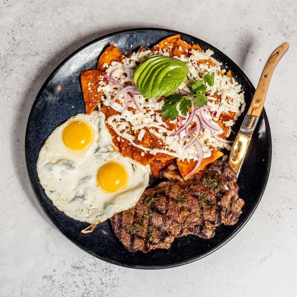 Overhead view of grilled steak with chimichurri, two fried eggs, saucy nachos, avocado, cotija cheese, red onion, and cilantro on a black plate.