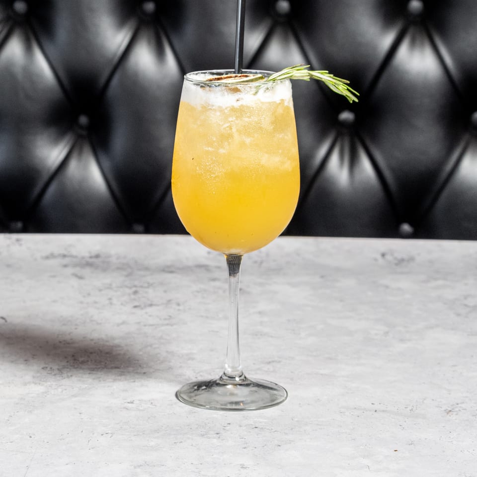 Golden cocktail in a stemmed glass, garnished with rosemary, sits on a marble bar against black tufted leather.