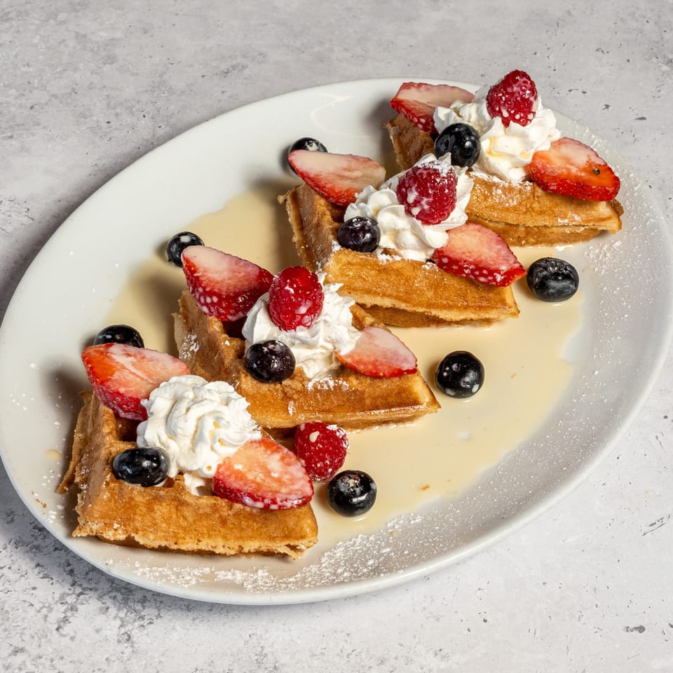 Golden waffles topped with whipped cream, strawberries, raspberries, blueberries, powdered sugar, and syrup on a white plate.